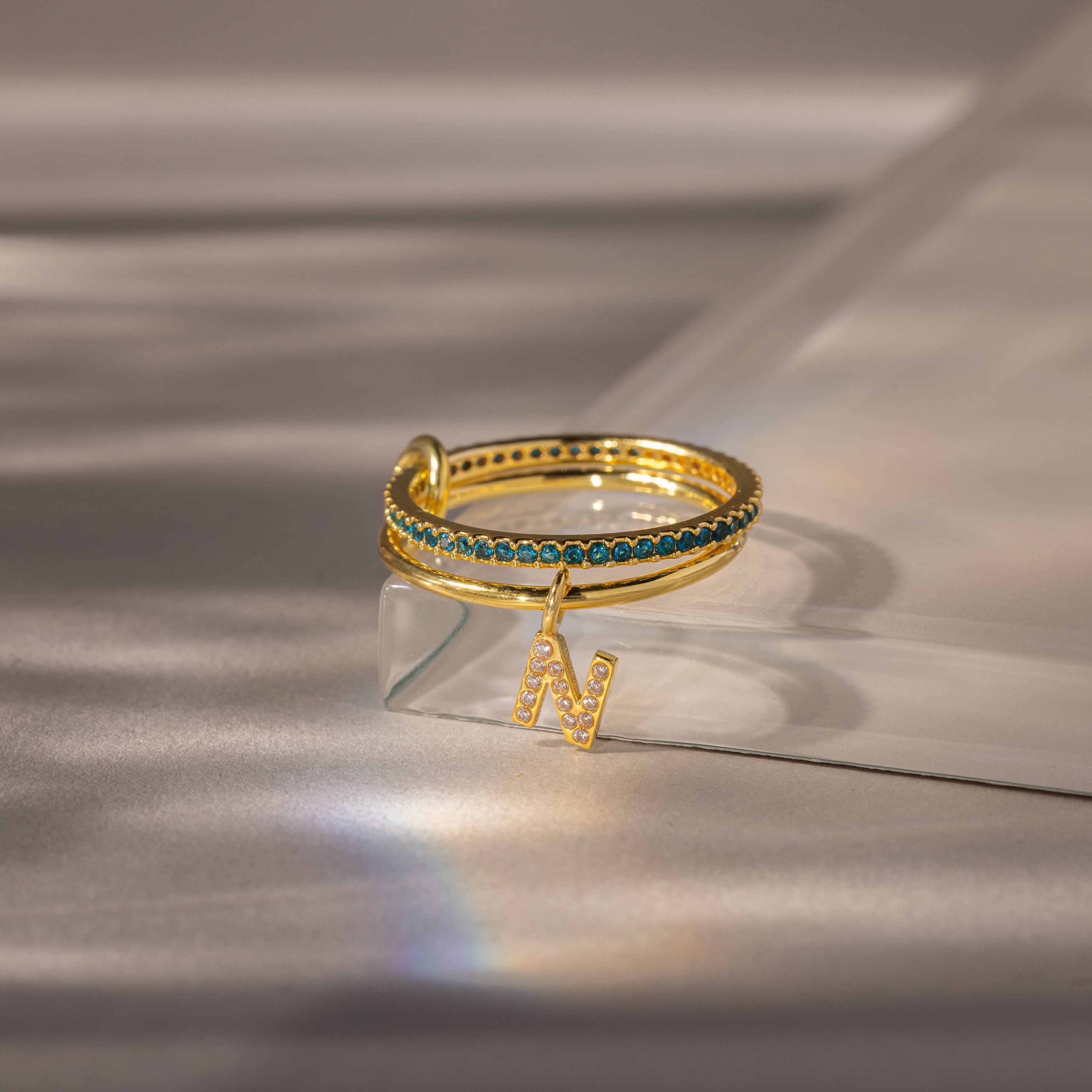 The Linked Initial Blue Zircon Ring Set features blue stones and a dangling letter N charm on a reflective surface—personalized jewelry designed to make your style unique.