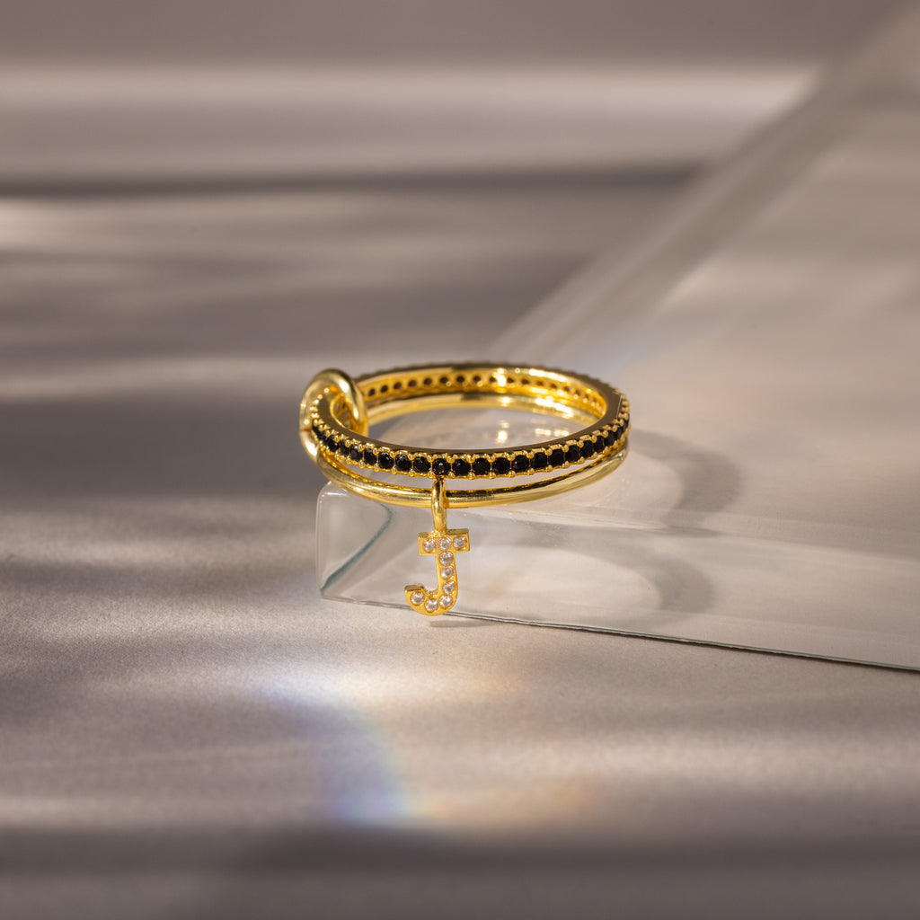 The Linked Initial Onyx Ring Set features a black stone accent and a dangling jeweled letter J charm, making it an elegant personalized jewelry piece or birthstone ring, beautifully displayed on a glass surface.