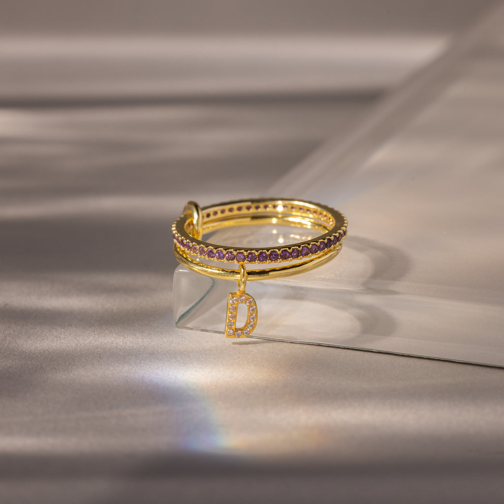 Linked Initial Birthstone Ring Set in Amethyst (February), featuring purple stones and a dangling letter D charm, displayed on a reflective surface.