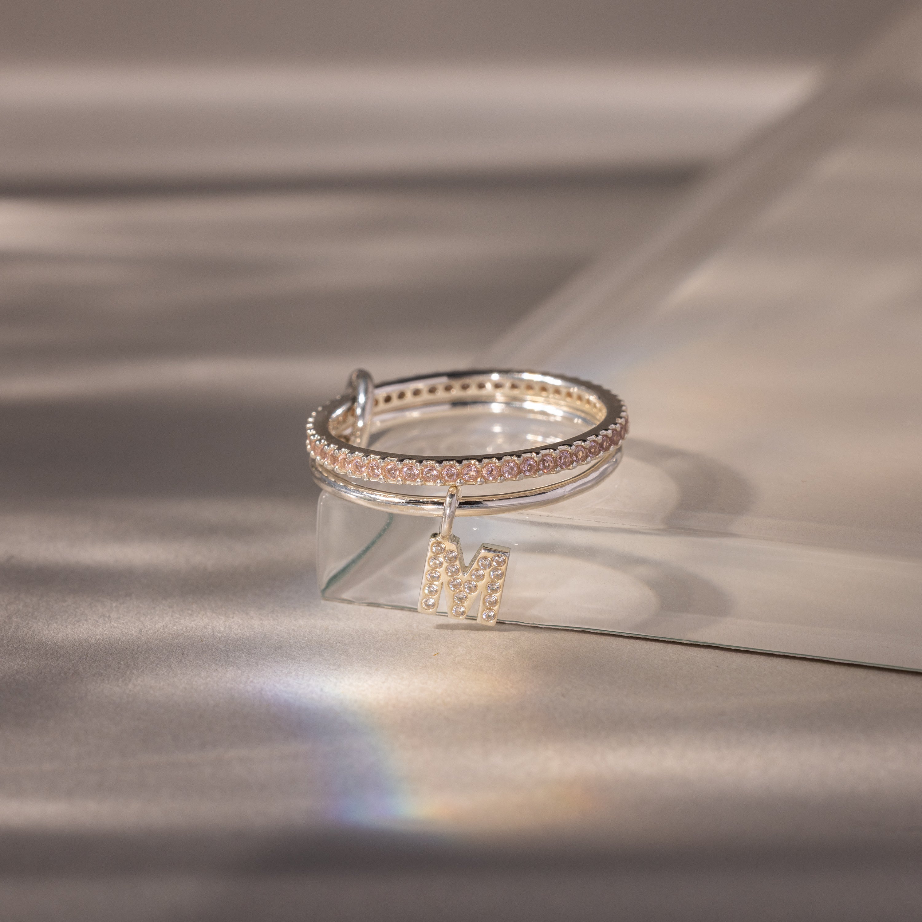 The Linked Initial Pink Tourmaline Ring Set features two silver rings with M charms and small gems, one accented by a delicate pink tourmaline birthstone, beautifully displayed on a clear, angled surface under soft lighting.