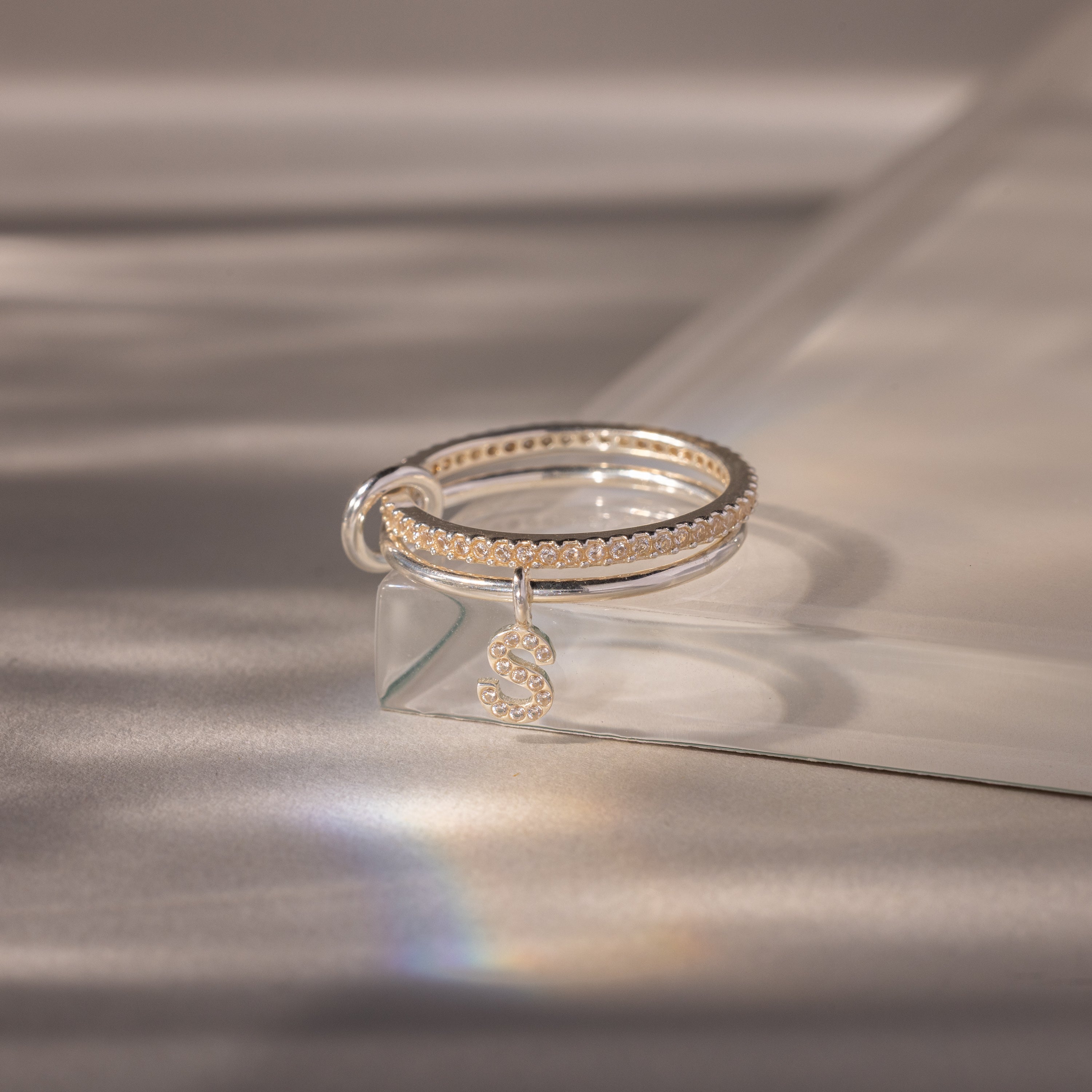 The Linked Initial Diamond Ring Set features a silver ring with a pavé letter S charm, adorned with small diamonds and elegantly displayed on a reflective surface.