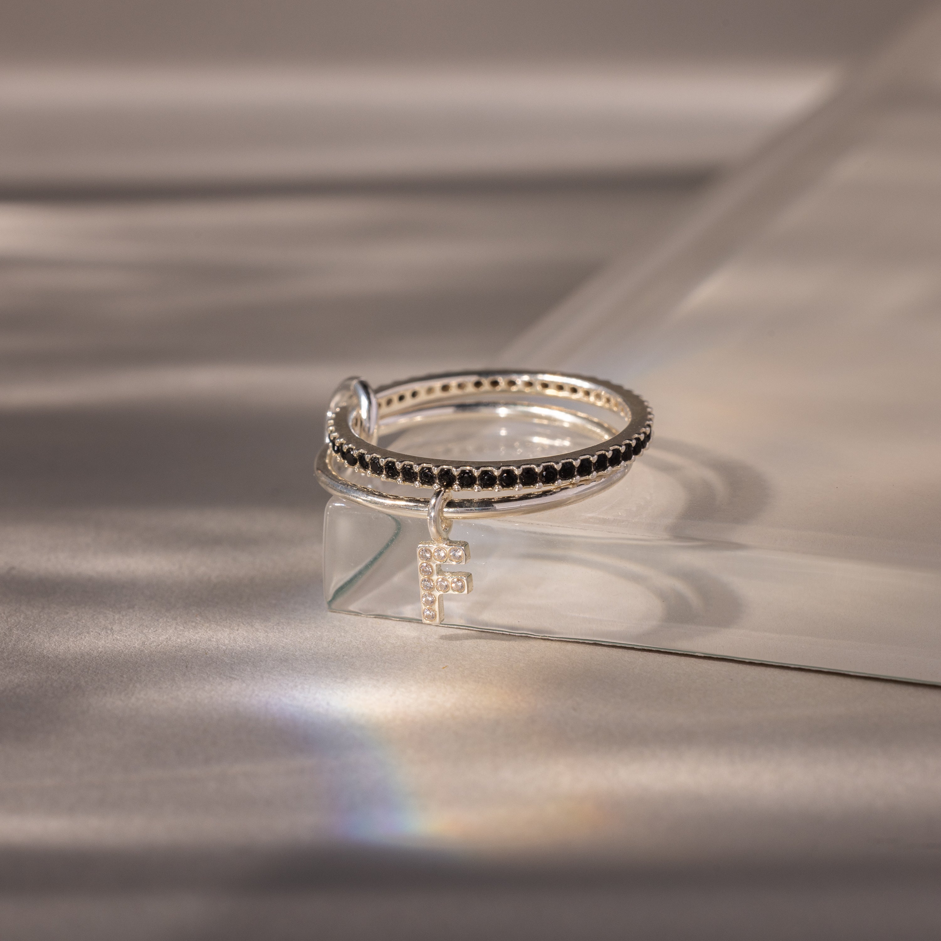 The Linked Initial Onyx Ring Set features black stones and a small cross charm, showcased on a reflective surface with soft lighting—a striking piece of personalized jewelry.