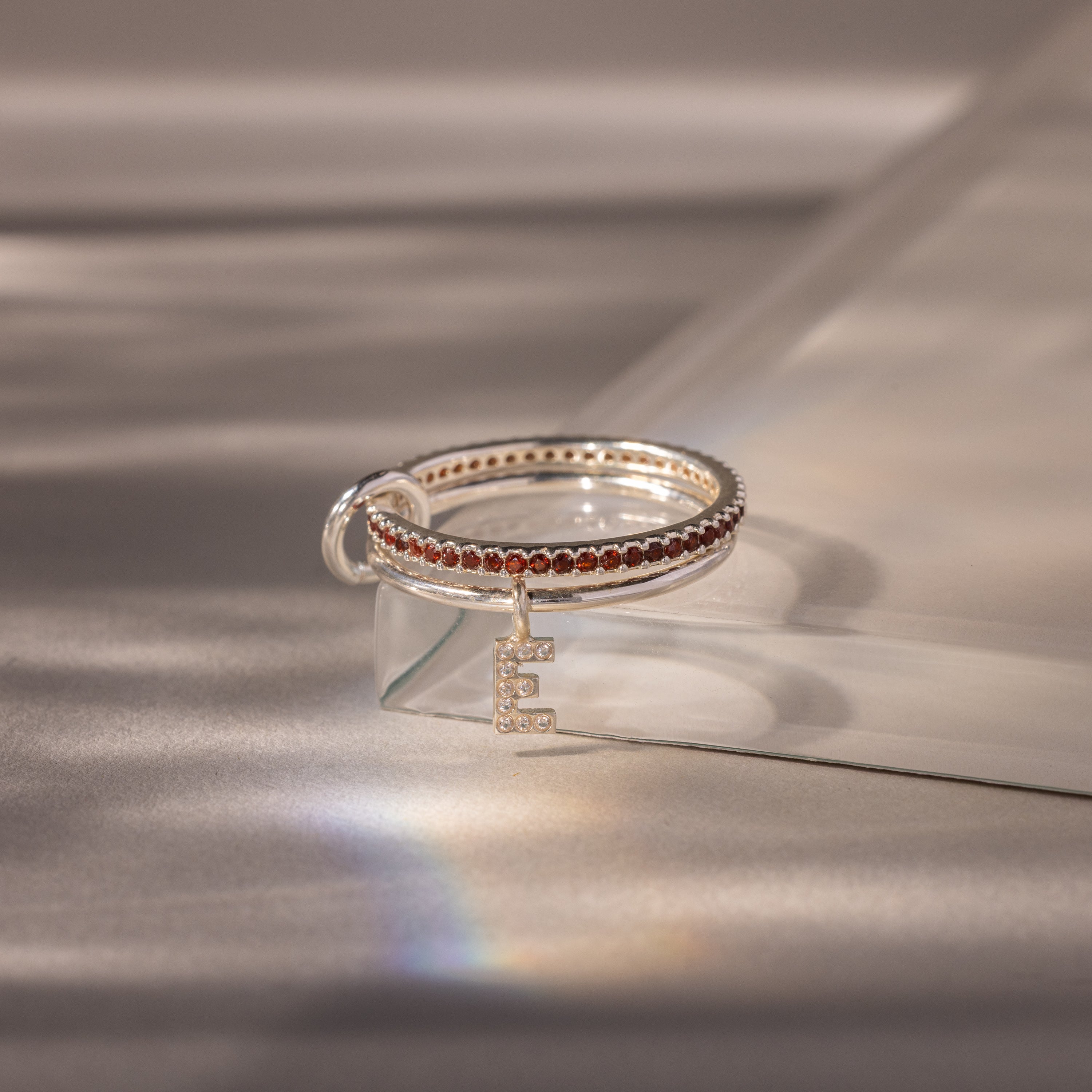 The Linked Initial Garnet Ring Set features red stones and a dangling letter E charm, shown on a glass block under soft lighting—a stylish and personalized jewelry piece.