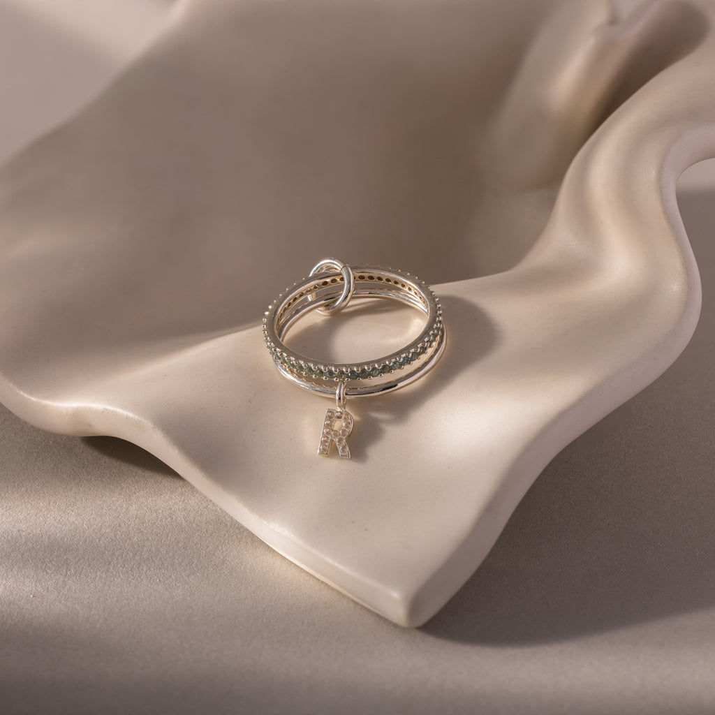 The Linked Initial Aquamarine Ring Set features silver rings with a small letter R charm, displayed on a smooth, wavy beige ceramic surface for an elegant personalized jewelry look.