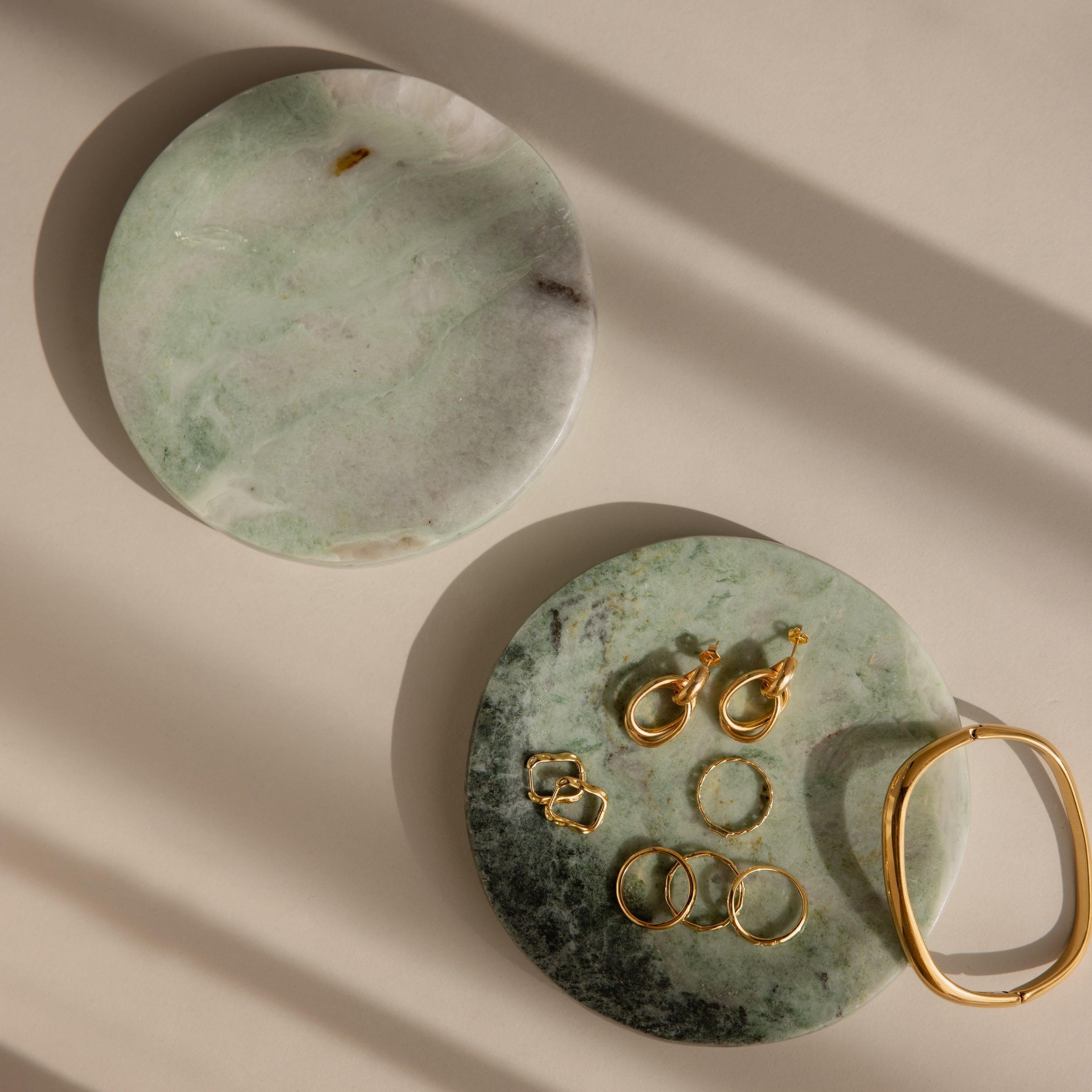 Two Green Marble Jewelry Dishes rest on a beige surface with soft shadows, one elegantly displaying gold jewelry.