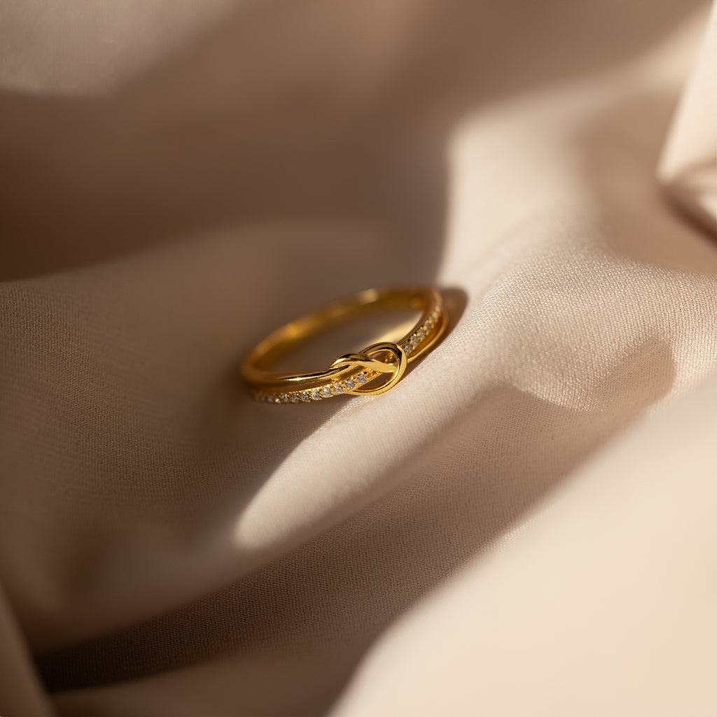 The Pave Knot Ring features an elegant knot design with sparkling accents—ideal as a jewelry gift—shown here resting on soft beige fabric in natural light.