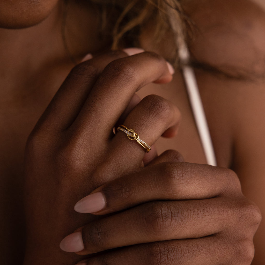 A manicured hand wears the Pave Knot Ring close to the chest—a gold ring featuring an elegant, timeless knot design and sparkling pavé, perfect as a jewelry gift.