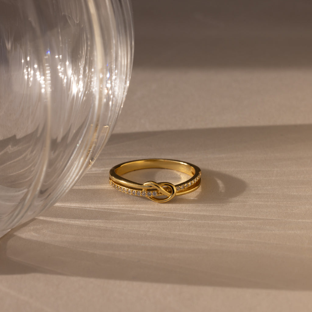 The Pave Knot Ring, adorned with small diamonds, is displayed next to a transparent glass on a beige surface.