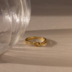 The Pave Knot Ring, adorned with small diamonds, is displayed next to a transparent glass on a beige surface.