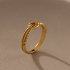 The Pave Knot Ring features gold plating with small embedded stones, showcased on a beige surface in soft lighting—an elegant and symbolic jewelry piece perfect for any occasion.