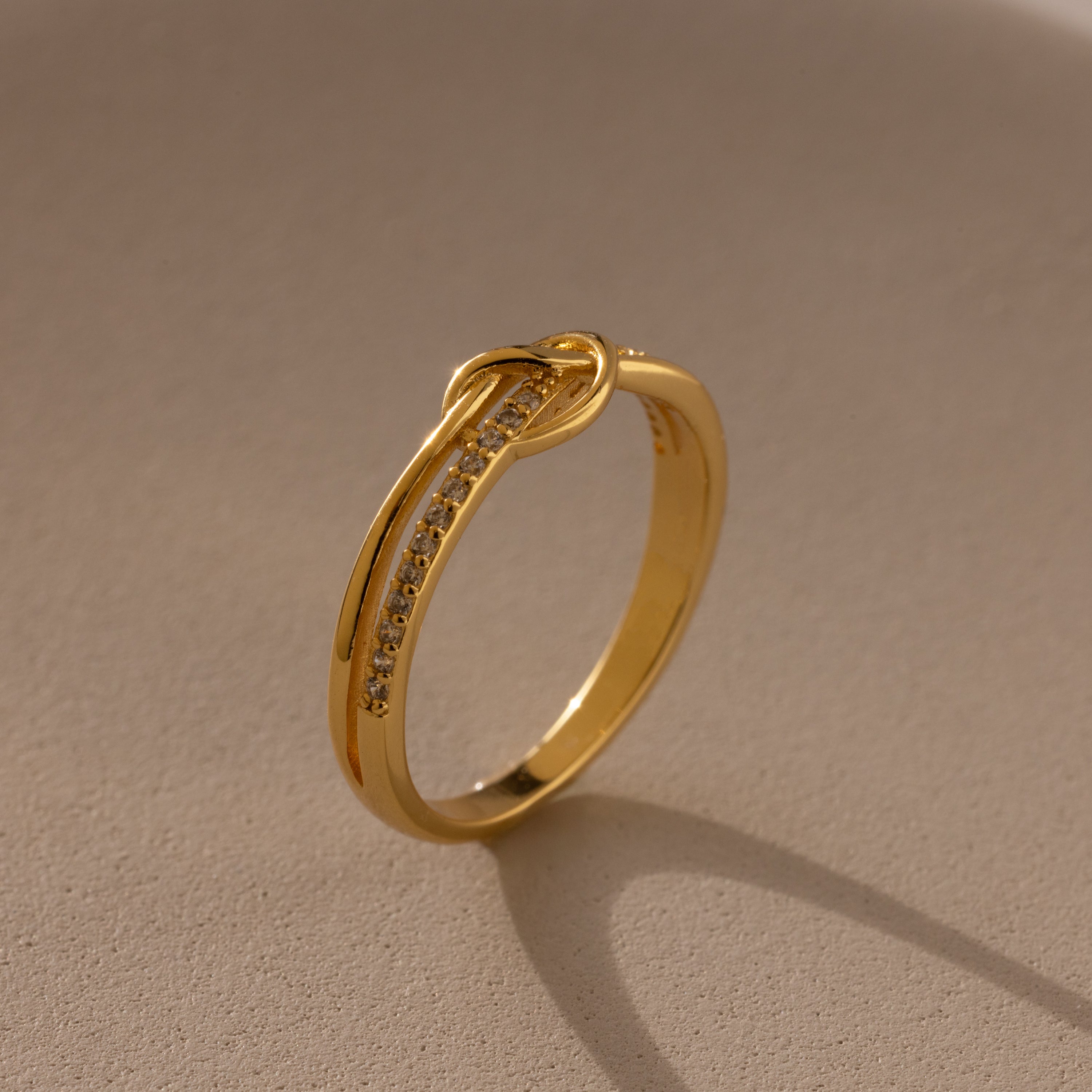 The Pave Knot Ring features gold plating with small embedded stones, showcased on a beige surface in soft lighting—an elegant and symbolic jewelry piece perfect for any occasion.