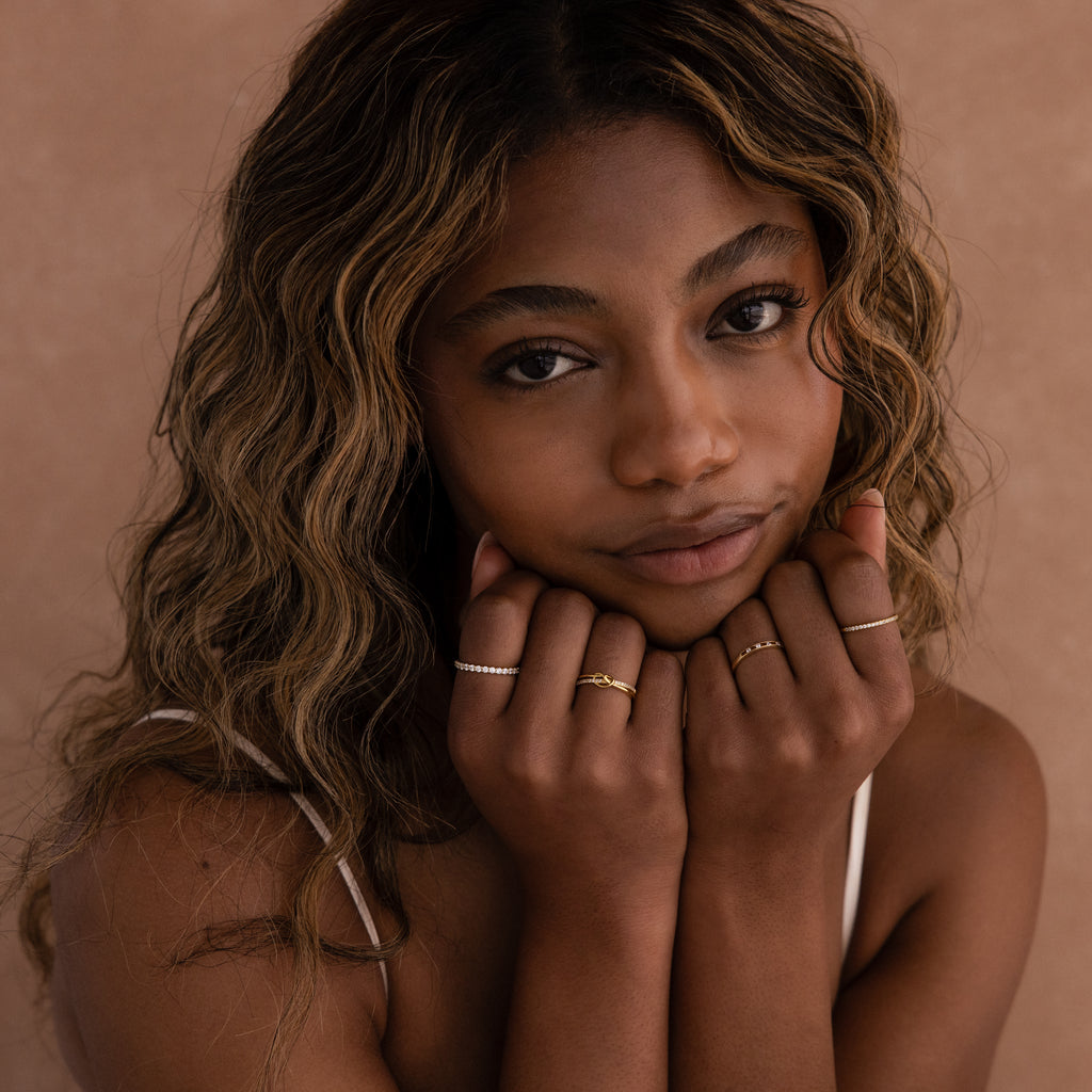 A woman with wavy hair poses against a beige background, showing off symbolic jewelry and multiple rings, including the Pave Knot Ring, with her hands near her face.