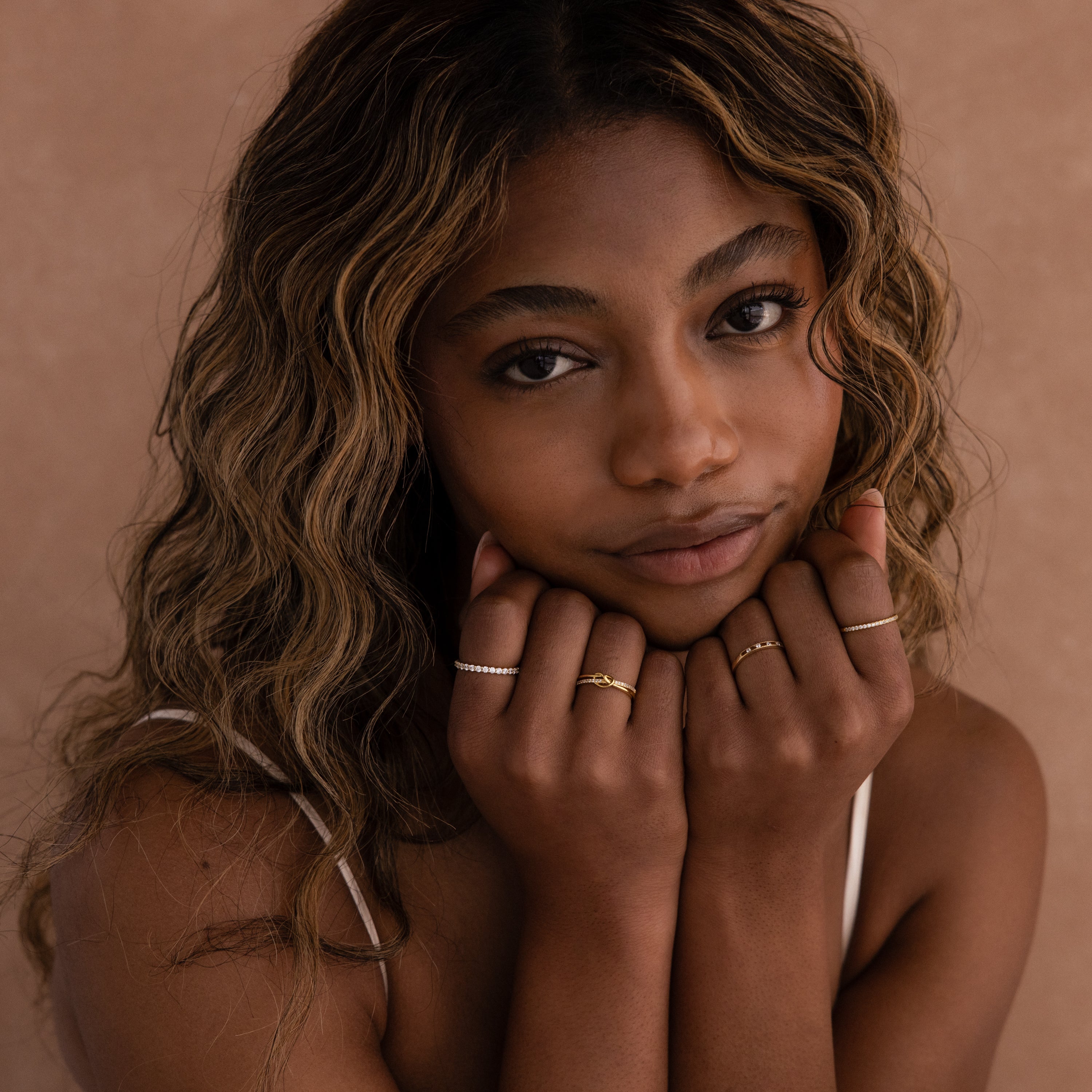 A woman with wavy hair poses against a beige background, showing off symbolic jewelry and multiple rings, including the Pave Knot Ring, with her hands near her face.