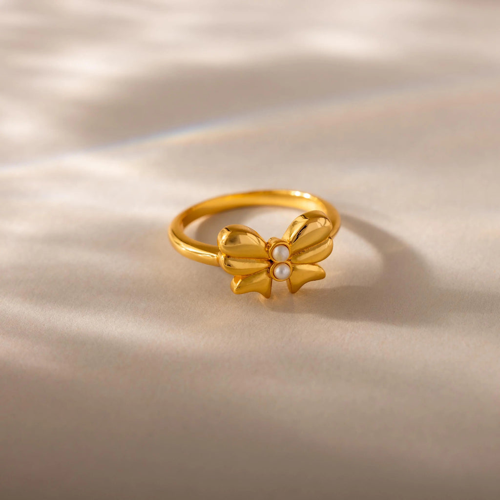 A close-up product shot of a gold ring featuring a prominent bow-shaped design at its center, set on a softly lit cream-colored surface. The bow is symmetrical and polished, with gracefully curved loops and pointed tails, evoking a delicate, feminine charm. At the center of the bow are two small, round white pearls stacked vertically.