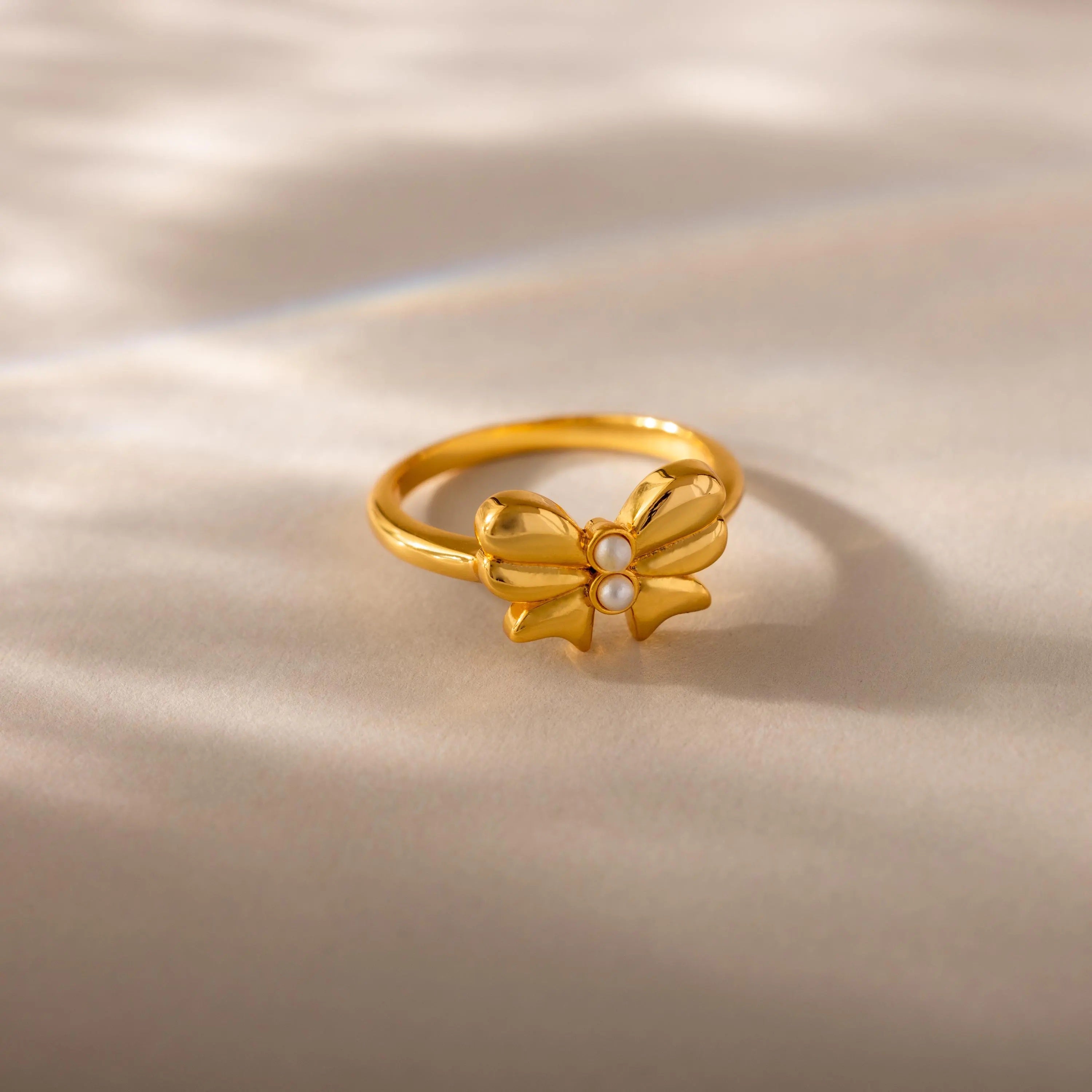 A close-up product shot of a gold ring featuring a prominent bow-shaped design at its center, set on a softly lit cream-colored surface. The bow is symmetrical and polished, with gracefully curved loops and pointed tails, evoking a delicate, feminine charm. At the center of the bow are two small, round white pearls stacked vertically.