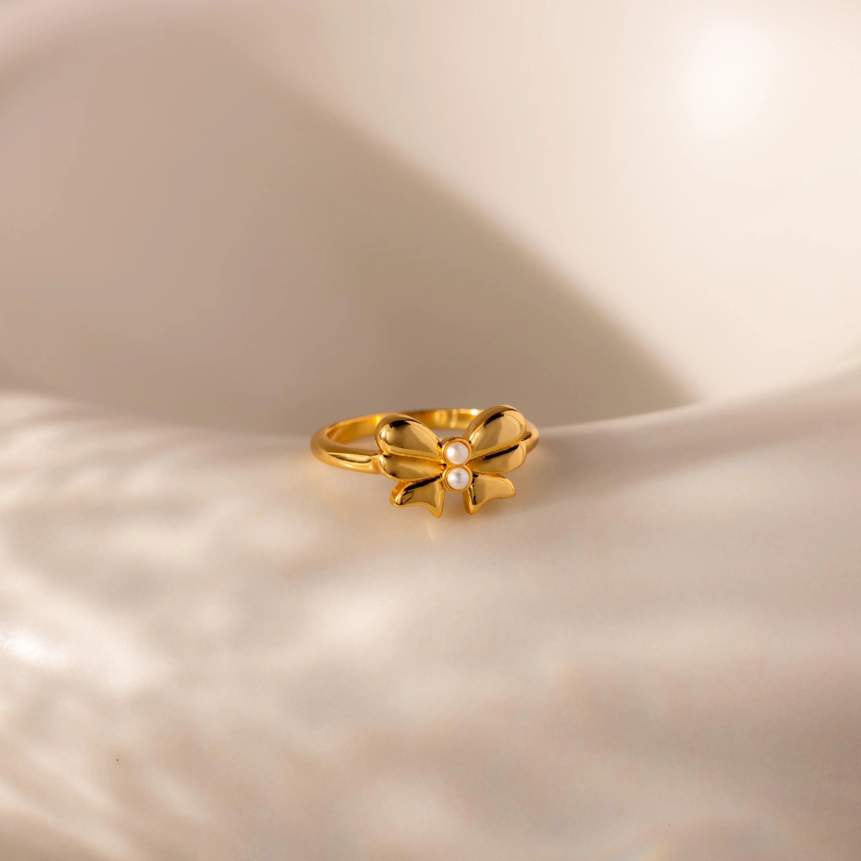 The gold Bow Pearl Ring is displayed on a softly lit, curved cream surface, highlighting its delicate design.