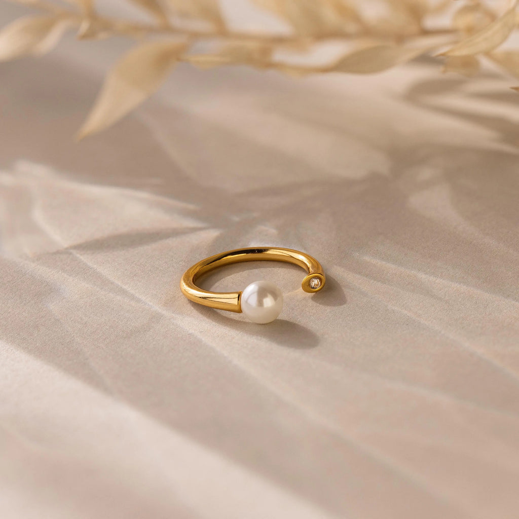 The Open Pearl & Diamond Ring, featuring a gold adjustable band with a pearl and small diamond, rests on a softly shadowed surface with dried leaves above.