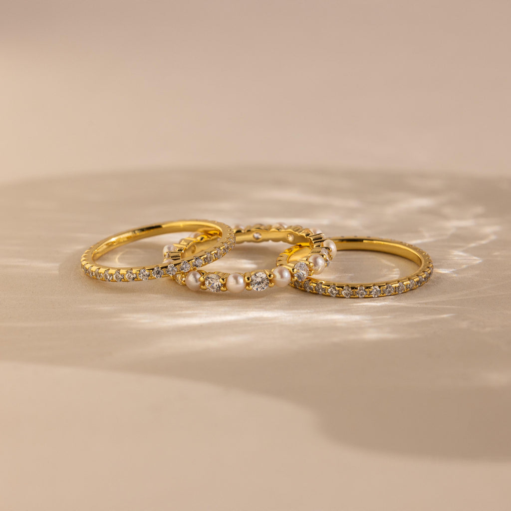 The Diamond Eternity Ring Set, featuring three gold rings, is displayed on a beige surface with soft lighting.