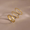 The Simple Ring Set features three thin gold stacking rings arranged in a row on a smooth beige surface with soft shadows, showcasing their elegant minimalist style.