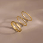 The Simple Ring Set features three thin gold stacking rings arranged in a row on a smooth beige surface with soft shadows, showcasing their elegant minimalist style.