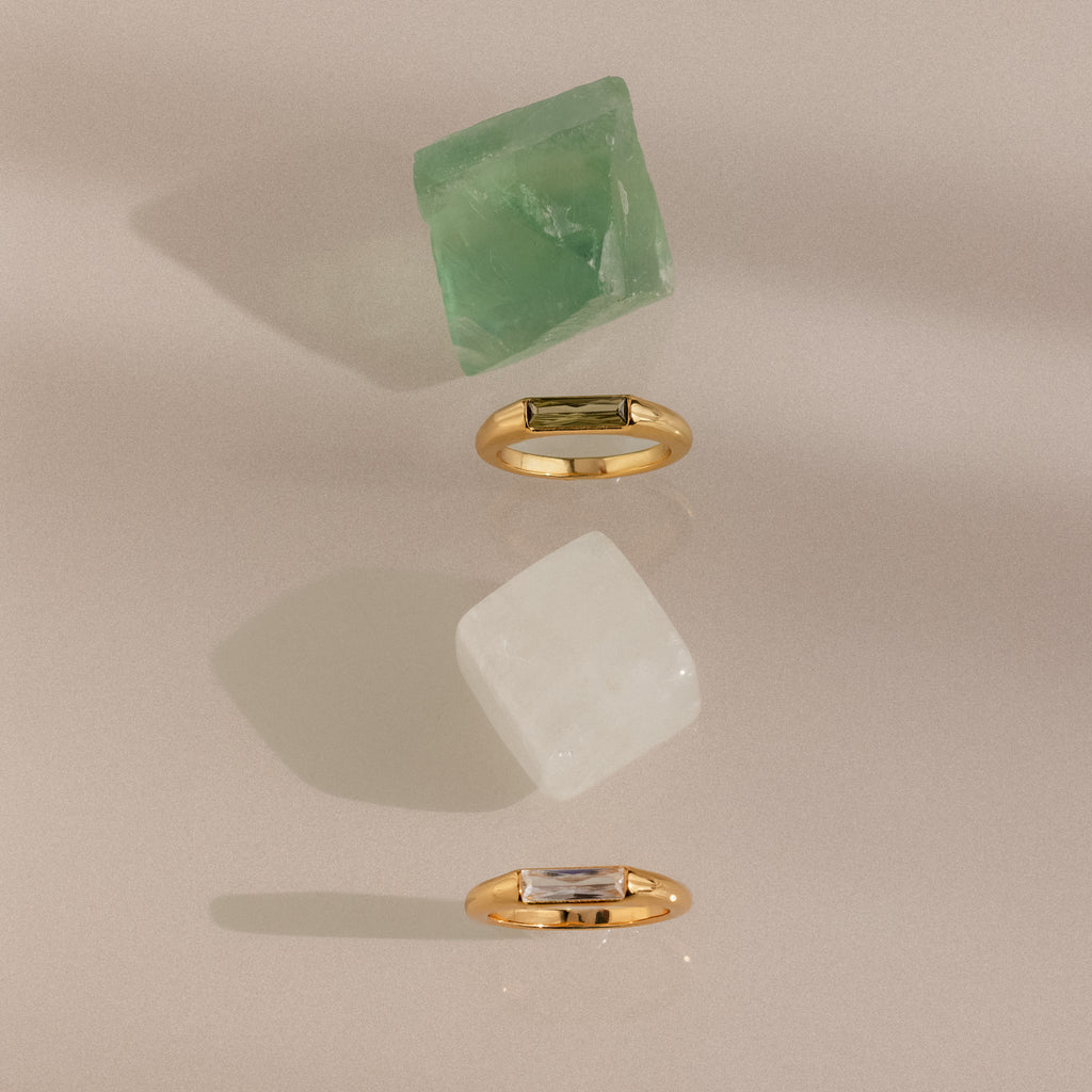 Two Baguette Signet Rings with rectangular gemstones are displayed beside green and white crystals on a neutral background, making a modern statement.