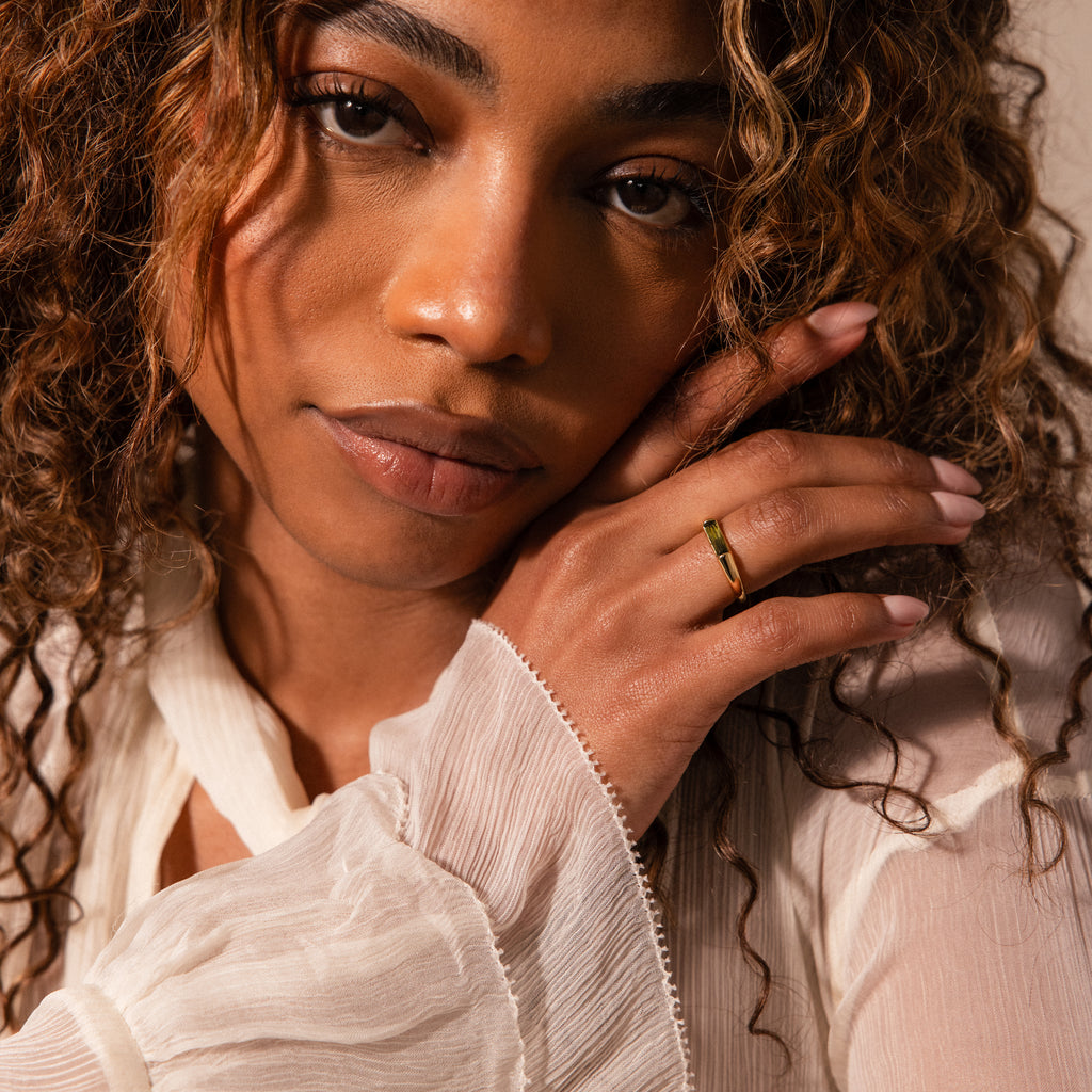 Woman with curly hair, wearing a light blouse and the Peridot Baguette Signet Ring, gently touches her face while looking at the camera.