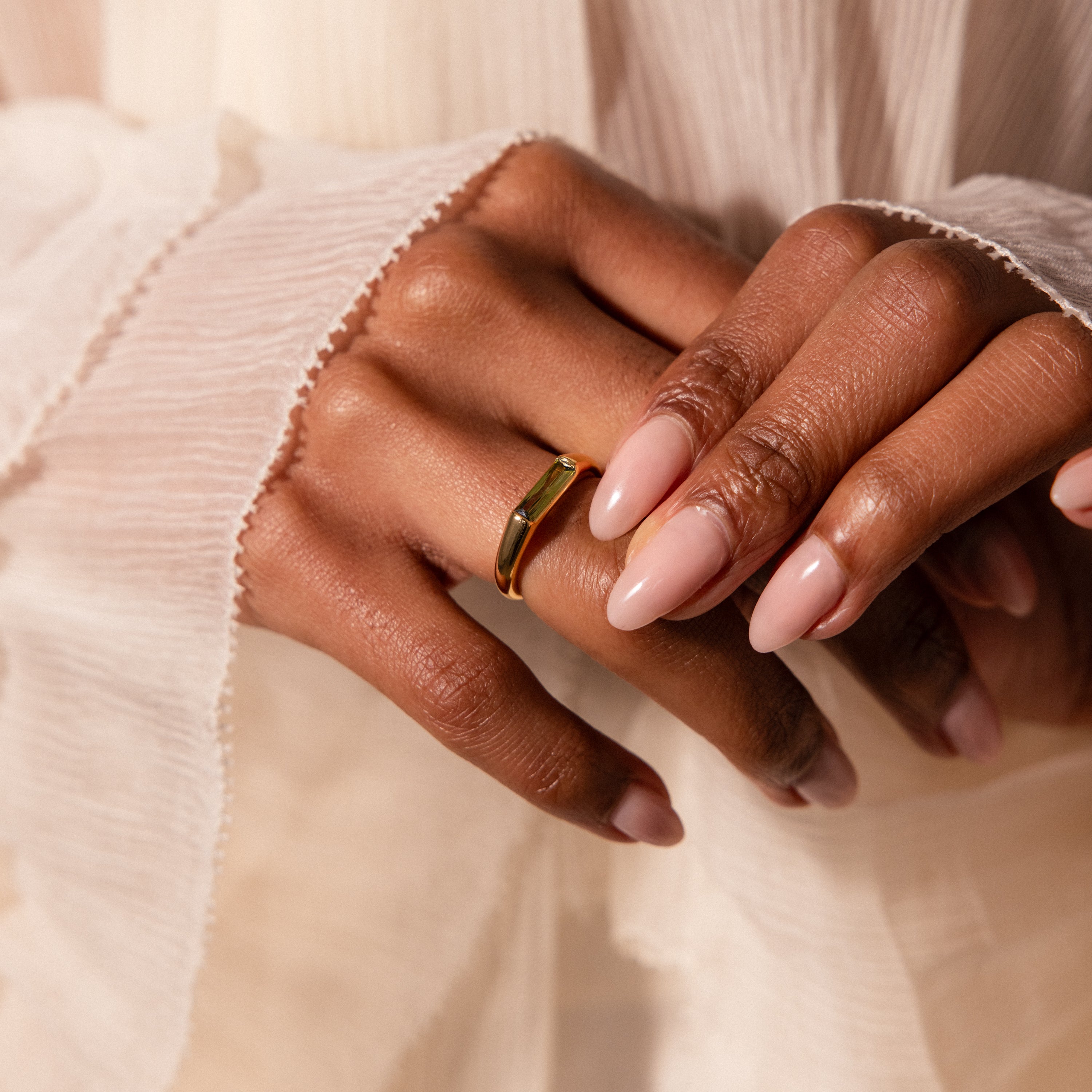 A hand with nude manicured nails wears the Peridot Baguette Signet Ring, resting on pleated cream fabric.