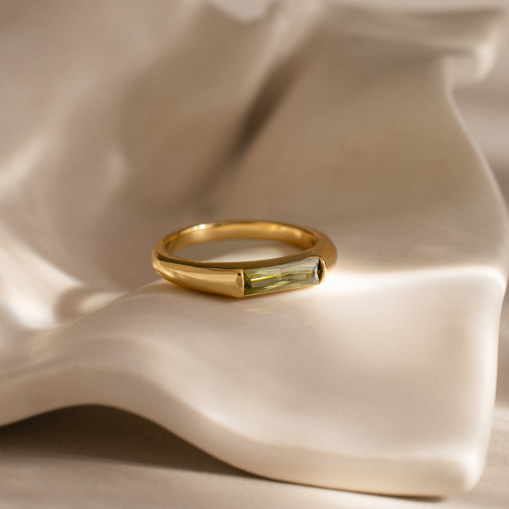 The Peridot Baguette Signet Ring, featuring a rectangular green gemstone set in gold, rests on a wavy cream ceramic surface, making a striking modern statement piece.