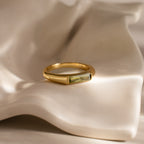 The Peridot Baguette Signet Ring, featuring a rectangular green gemstone set in gold, rests on a wavy cream ceramic surface, making a striking modern statement piece.