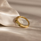 The Peridot Baguette Signet Ring, a modern gold statement piece with a green gemstone, rests on a smooth white surface under soft lighting.