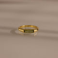 The Peridot Baguette Signet Ring is a modern gold statement ring featuring a rectangular green peridot gemstone in a baguette cut, showcased on a smooth beige surface with soft shadows.