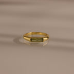 The Peridot Baguette Signet Ring is a modern gold statement ring featuring a rectangular green peridot gemstone in a baguette cut, showcased on a smooth beige surface with soft shadows.