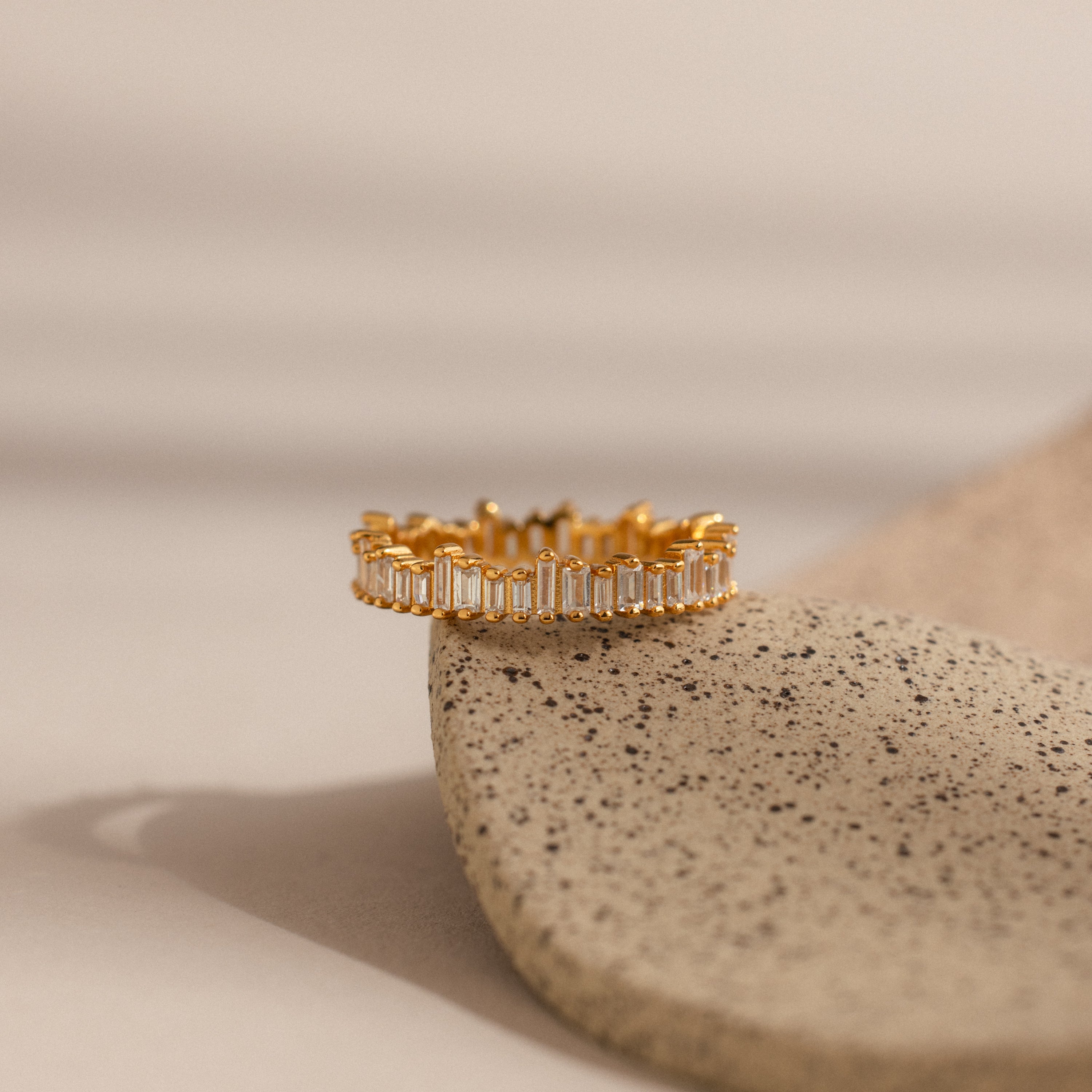 The Art Deco Rhythm Ring, featuring rectangular baguette diamonds, is displayed on a speckled beige stone against a soft neutral backdrop.