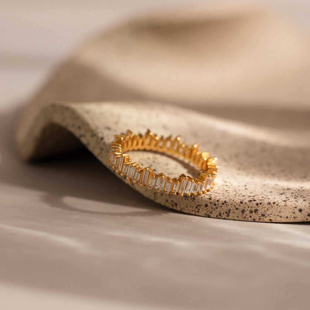 The Art Deco Rhythm Ring, designed in gold with rectangular baguette diamonds and made to stack, rests on a curved, textured beige surface.