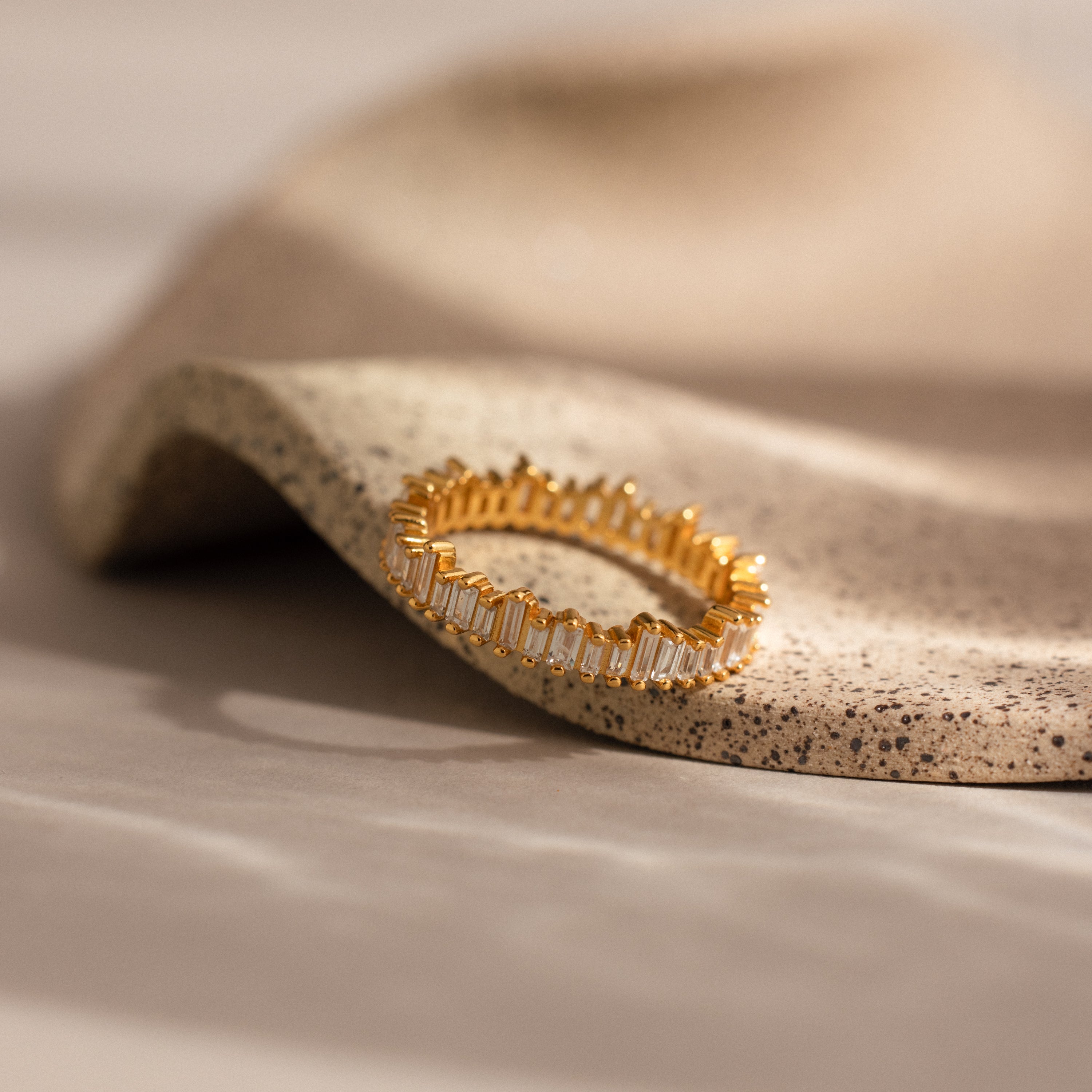 The Art Deco Rhythm Ring, designed in gold with rectangular baguette diamonds and made to stack, rests on a curved, textured beige surface.