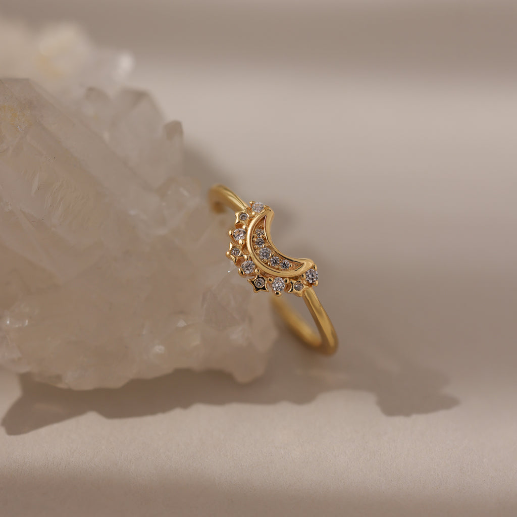The Diamond Crescent Moon Ring, featuring a gold band with a crescent moon and small diamonds, is displayed on a clear crystal against a neutral background.