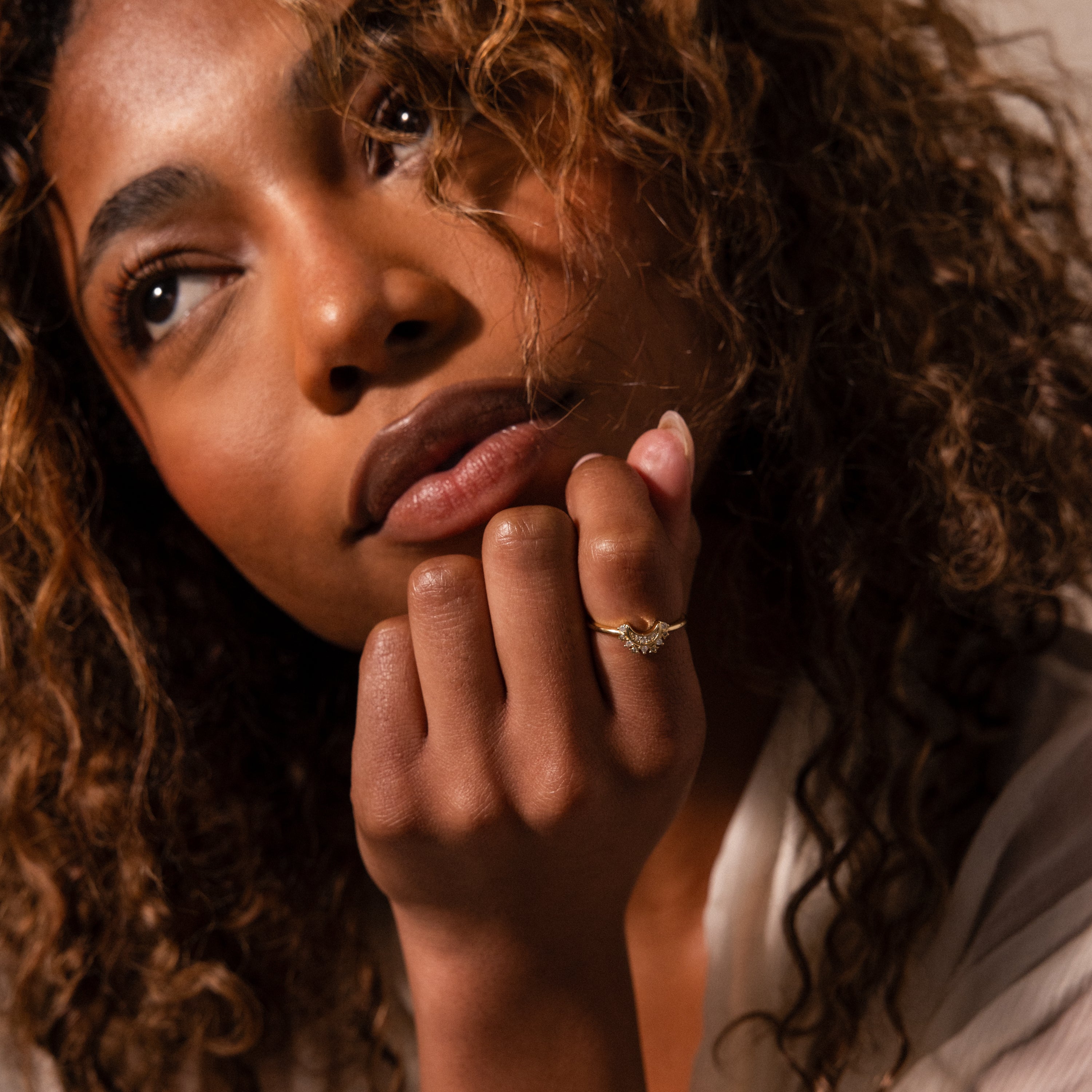 A woman with curly hair gazes thoughtfully, displaying the Diamond Crescent Moon Ring on her hand near her chin.