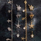 The Dainty Shark Tooth Charm in 18K Gold shines among ocean-themed bracelets and charms—turtles, hearts, starfish, and pearls—perfect coastal jewelry displayed on a dark surface.