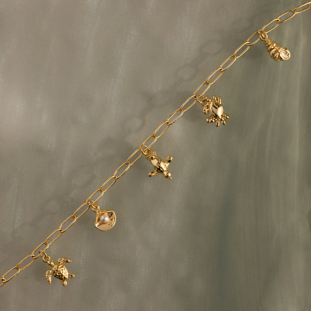 Seaside Charm Bracelet in 18K Gold: Features ocean-inspired turtle, shell, fish, and seahorse charms—ideal for coastal jewelry lovers. Shown on a gray background.