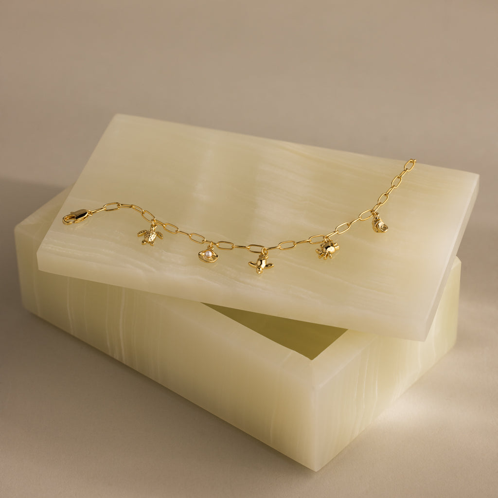 The Seaside Charm Bracelet in 18K Gold features ocean-inspired turtle, crab, starfish, and seashell charms, elegantly displayed on a marble box—perfect for lovers of coastal jewelry.
