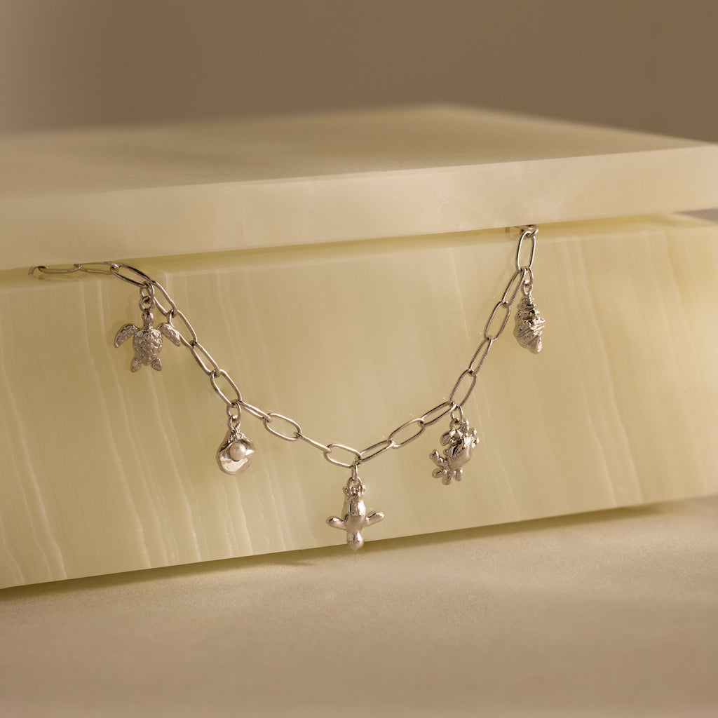 The Seaside Charm Bracelet features turtle, moon, cross, and pineapple ocean-inspired charms displayed on a cream-colored stone box.