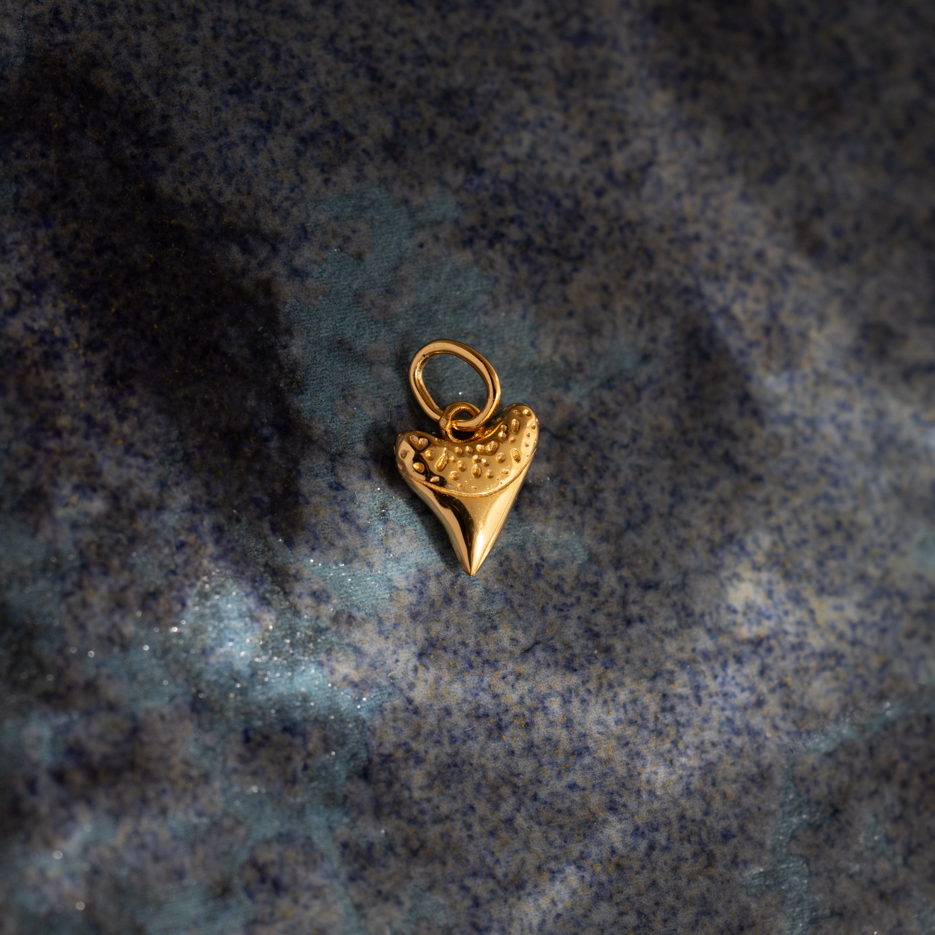 The Dainty Shark Tooth Charm in 18K Gold rests on a blue marbled surface, making it an ideal accessory for coastal jewelry enthusiasts.