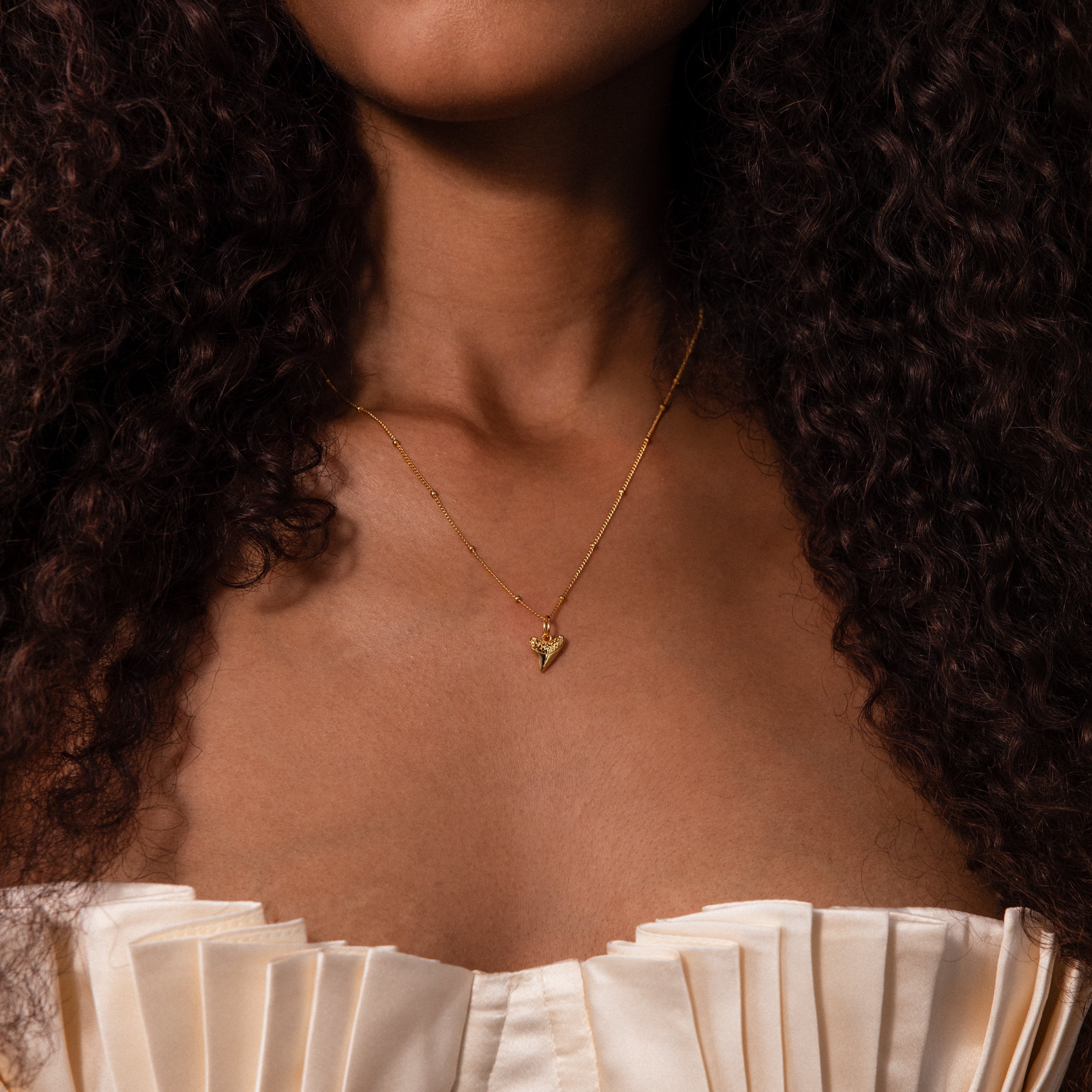 Woman wearing the Shark Tooth Necklace in 18K Gold, featuring an Africa-shaped pendant, paired with a cream pleated off-shoulder top.
