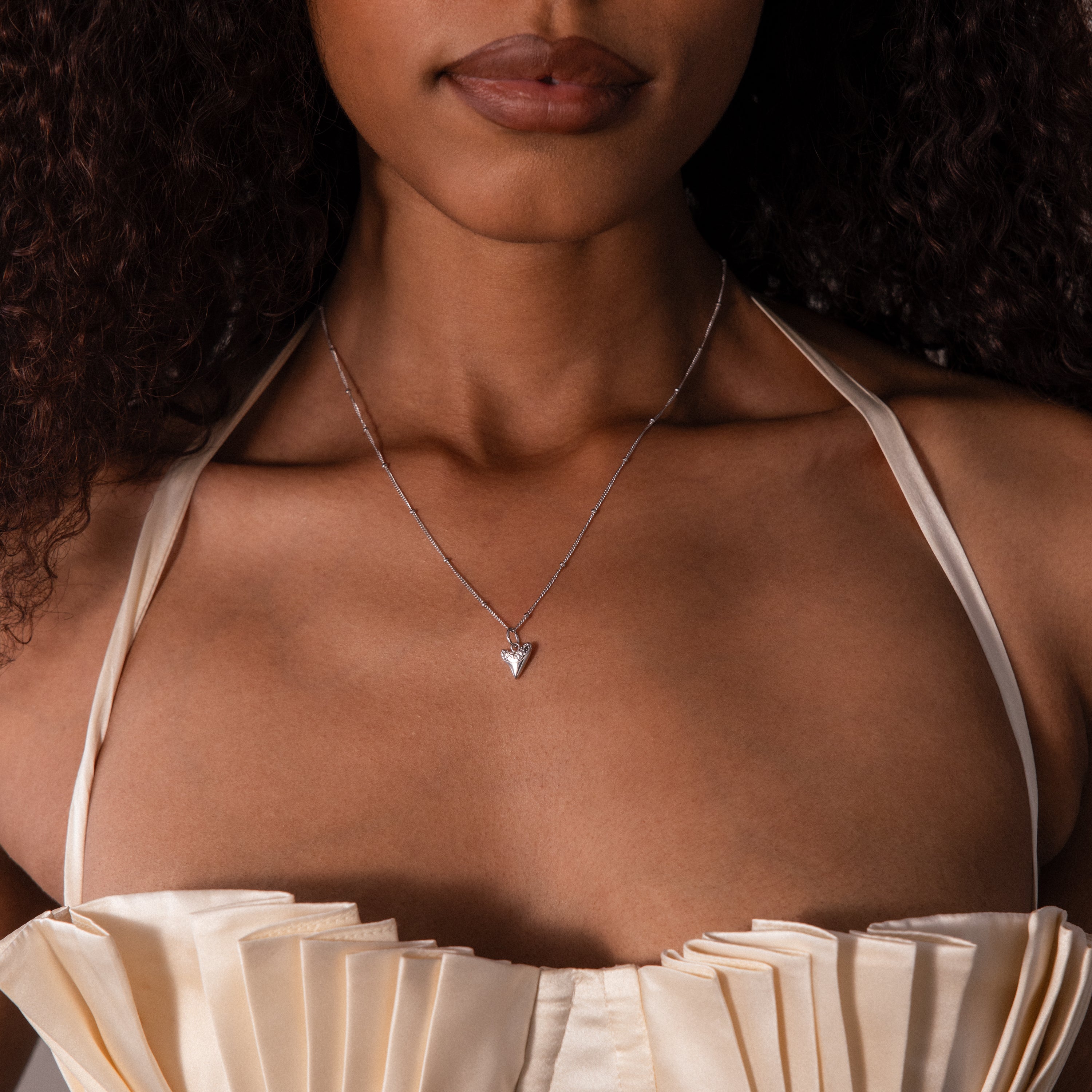 A woman wears a cream pleated top and the Shark Tooth Necklace, featuring a silver triangle pendant, for a chic beachside look.