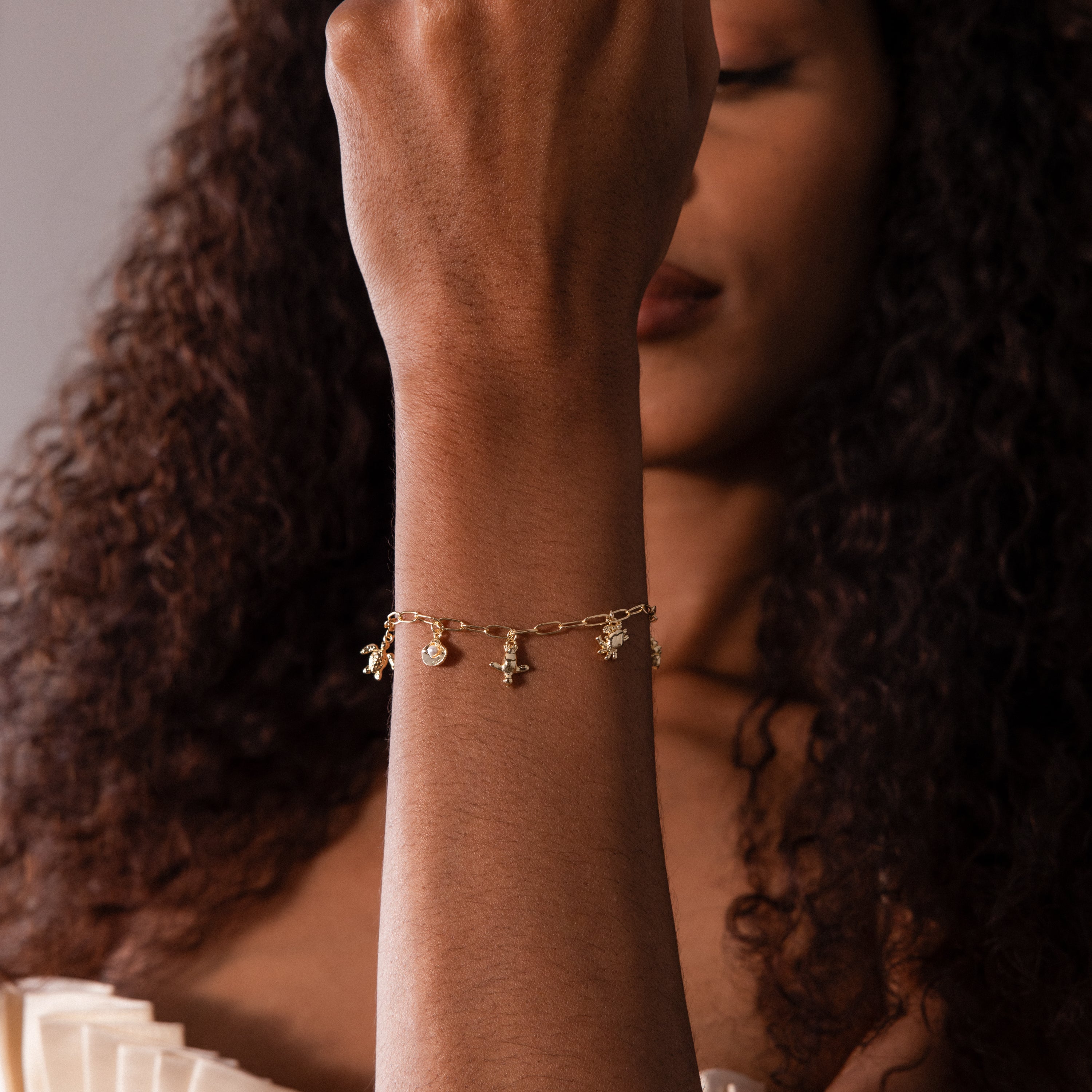 A woman with curly hair in a cream off-shoulder top raises her arm, displaying the Seaside Charm Bracelet in 18K Gold adorned with ocean-inspired charms.