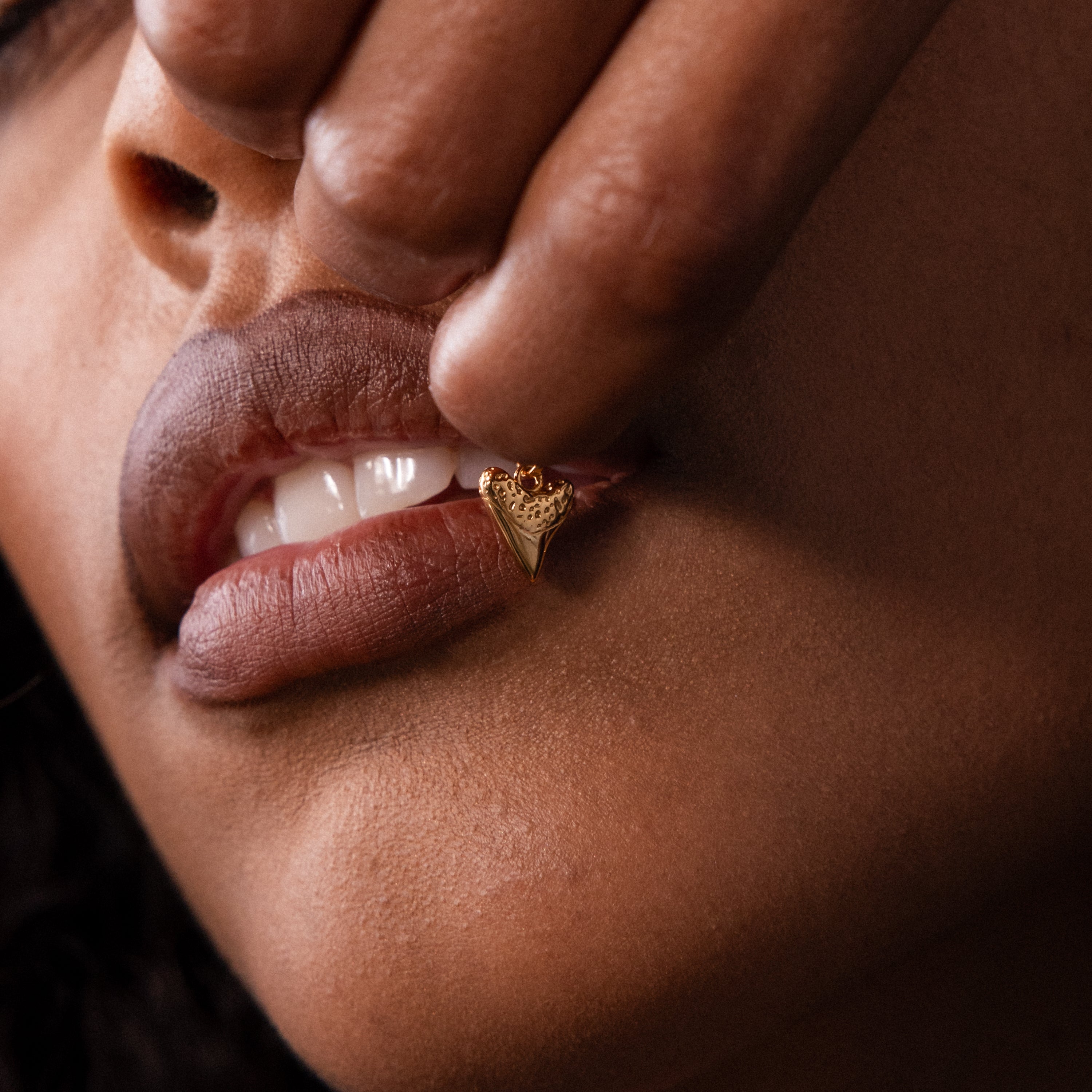 Close-up of a person biting their lip, holding the Dainty Shark Tooth Charm in 18K Gold near their mouth, showcasing trendy coastal jewelry.