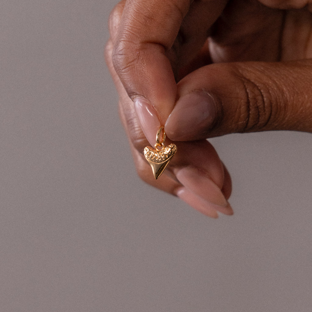 A hand holds the Dainty Shark Tooth Charm in 18K Gold, a pointed heart-shaped pendant with tiny stones at the top—perfect for lovers of coastal jewelry.