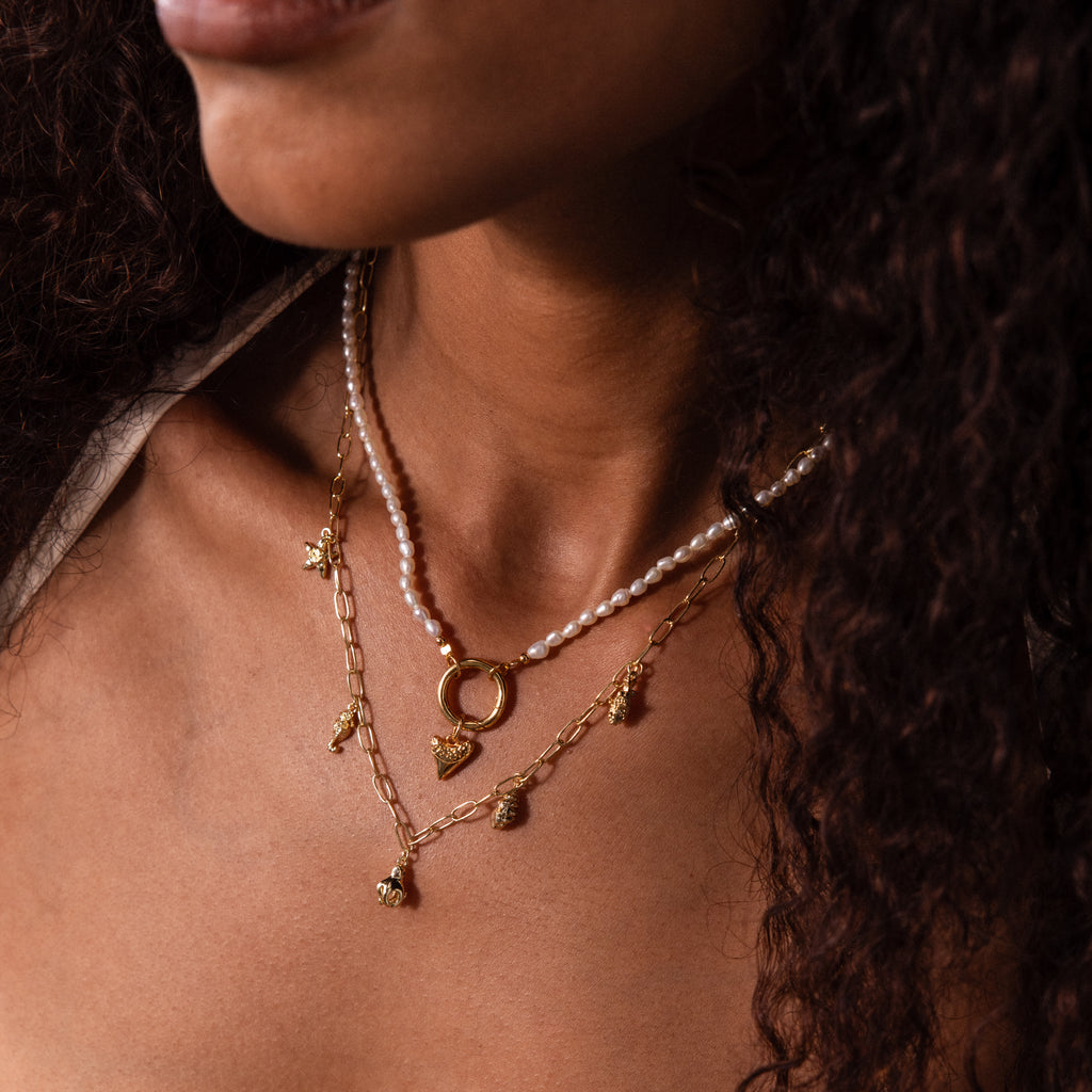 Close-up of a woman with curly hair wearing layered gold and pearl necklaces, featuring coastal jewelry and the Dainty Shark Tooth Charm in 18K Gold.