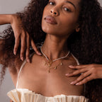 Woman with curly hair wearing layered Coastal Jewelry, featuring a silver Dainty Shark Tooth Charm necklace and a cream pleated top, posing with hands near her face.