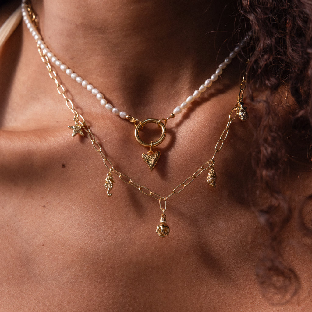 Close-up of layered gold necklaces with charms, a pearl strand, and the Dainty Shark Tooth Charm in 18K Gold, creating an elegant coastal jewelry look on the neck and collarbone.